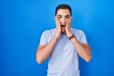 Young hispanic man standing over blue background afraid and shocked, surprise and amazed expression with hands on face 