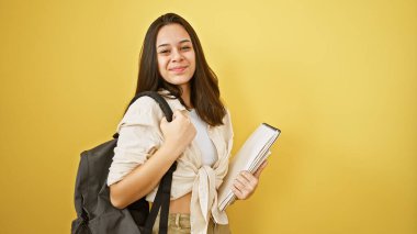 Gülümseyen, genç ve güzel İspanyol bir kadın öğrenci. Kendine güveni tam, sırt çantası ve kitabı olan, izole edilmiş sarı bir arka planda poz veren, eğlenceli ve pozitif bir yaşam tarzının tadını çıkaran.