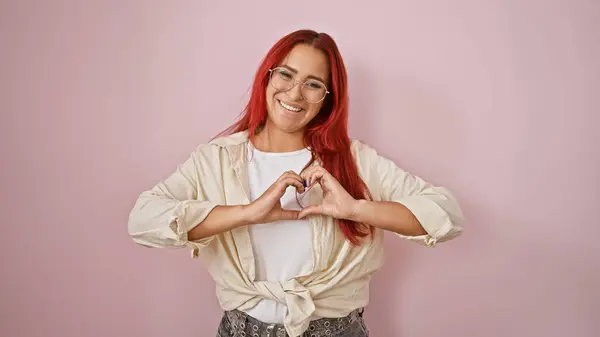 陽気な若い赤毛の女性は 自信を持って微笑み 孤立したピンクの背景の上に手で愛の心をジェスチャー — ストック写真