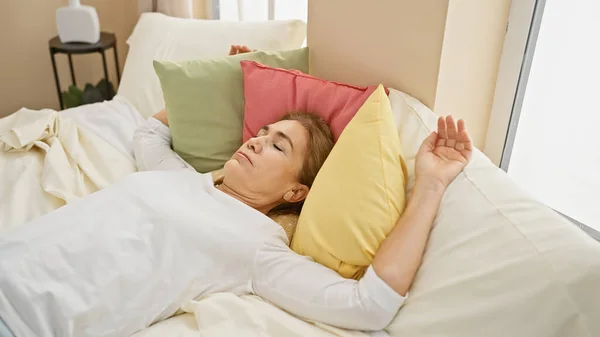 Mature blonde woman sleeping peacefully in a cozy bedroom with colorful pillows, depicting rest and comfort.