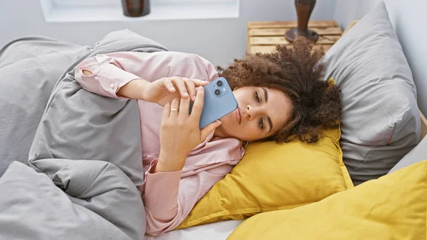 一个年轻的惊慌失措的女人 卷曲的头发躺在床上 用她的智能手机在室内 — 图库照片