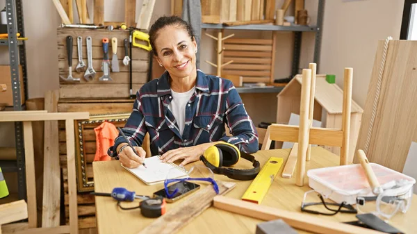 Smiling mature hispanic woman drafting in a woodwork studio surrounded by tools and wooden objects.