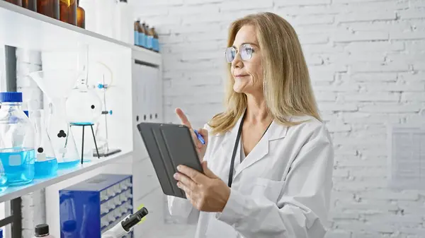 A mature woman scientist analyzes data on a tablet in a white laboratory with various chemicals and equipment.