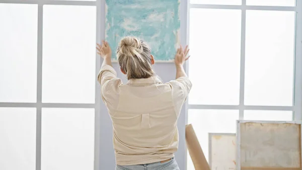 Young blonde woman artist hanging draw on wall at art studio