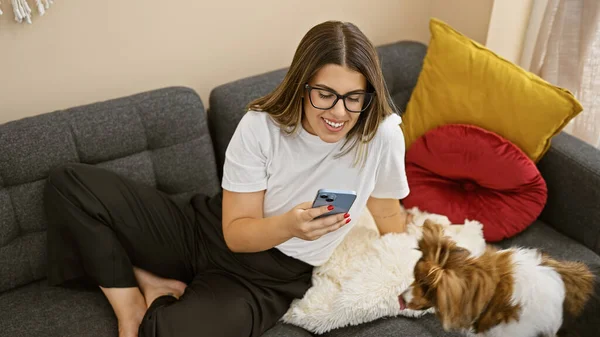 一位戴着眼镜 面带微笑的年轻女子和她的狗坐在沙发上 在舒适的客厅里用智能手机 — 图库照片