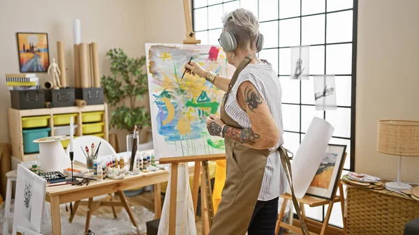Passionate grey-haired senior woman artist, indulged in drawing while listening to soothing music in her cozy art studio