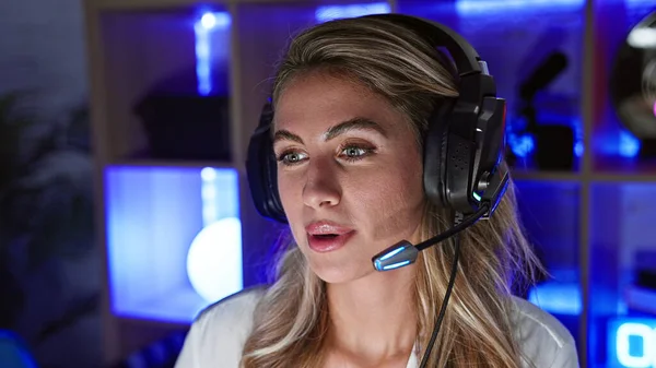 Focused blonde streamer, young woman with serious face gaming, headphones on in dark gaming room, living the futuristic cyber video stream life