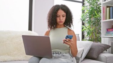 Çekici, kıvırcık saçlı, evine odaklanmış, dizüstü bilgisayar ve akıllı telefon kullanan, teknoloji dünyasına dalmış genç İspanyol bir kadın..