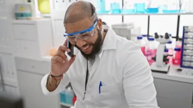 Laboratuvar önlüklü, sakallı, laboratuvar ortamında telefonla konuşan mutlu Afrikalı Amerikalı adam.