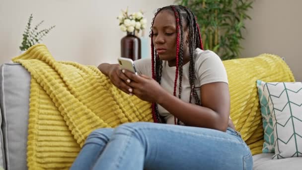 身穿辫子坐在家里沙发上 沉浸在客厅里用智能手机的非洲裔美国女人很不安 — 图库视频影像