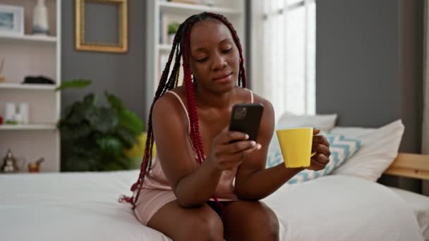 アフリカ系アメリカ人女性 ベッドで快適に目を覚まし コーヒーを飲んで かわいいアパートのベッドルームでスマートフォンにテキストを送っている間 すべての笑顔 — ストック動画