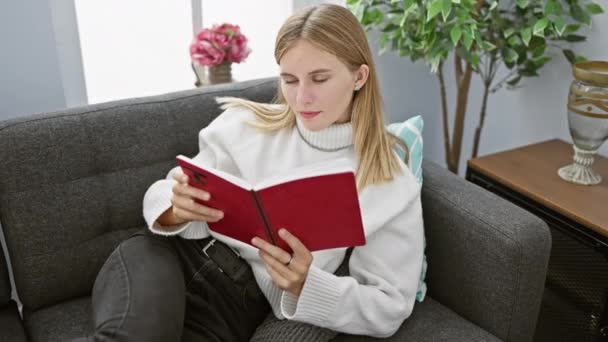 Μια Νεαρή Ξανθιά Γυναίκα Απολαμβάνει Την Ανάγνωση Στο Σύγχρονο Σαλόνι — Αρχείο Βίντεο