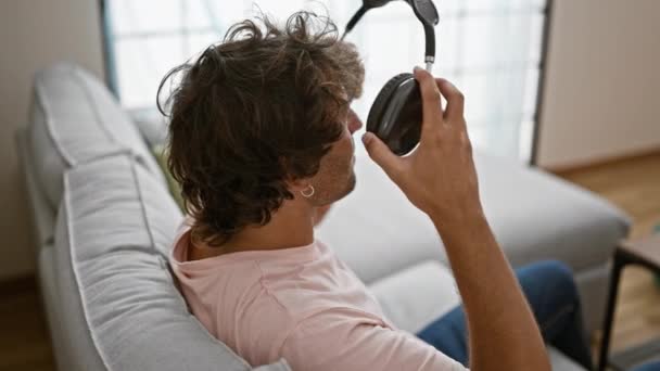 在明亮 现代的客厅里 一个放松的男人躺在沙发上 一边用耳机听音乐 — 图库视频影像
