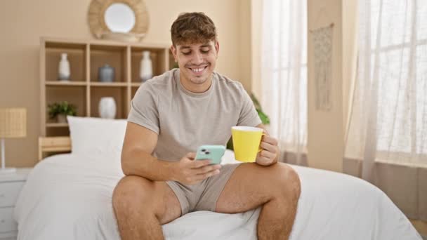 英俊的年轻人在智能手机上愉快地发短信 在床上喝咖啡时放松 体现了舒适 充满活力的卧室里完美舒适的室内环境 — 图库视频影像