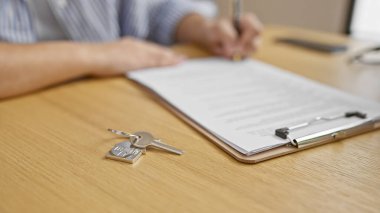 Yakın plan bir fotoğraf, bir emlak anlaşmasını resmetmek için evin anahtarlarının yanında, ahşap bir masada belgeleri imzalayan bir adamı yakalıyor..