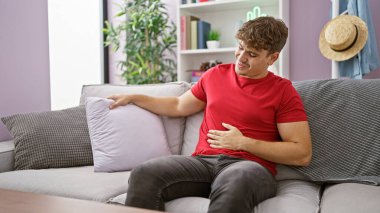 Ciddi karın ağrısı çeken endişeli genç bir İspanyol, evinde oturma odasındaki bir kanepede dinlenirken mutsuz bir şekilde karnına dokunuyor. Kapalı alan hastalığının resmini çiziyor..