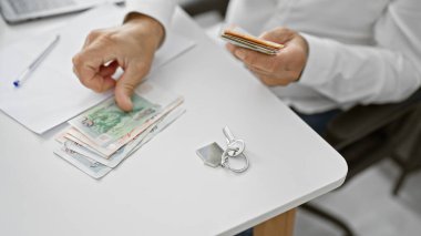 Ofiste bir adam dizüstü bilgisayar ve anahtarların yanında para sayıyor, iş ve finansmanı simgeliyor..