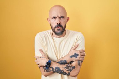 Sarı arka planda dikilen İspanyol dövmeli adam. Kış soğuğu yüzünden titriyor ve donuyor. Yüzünde üzgün ve şok ifadesi var. 