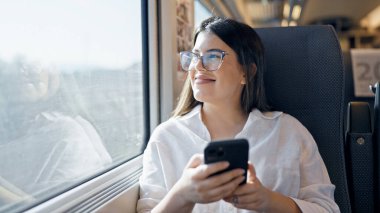 Genç, güzel, İspanyol bir kadın gülümsüyor. Akıllı telefon kullanıyor. Trende oturuyor.