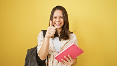 Genç ve güzel bir İspanyol kadının neşeli portresi, güneşli sarı bir arka planda izole bir şekilde okul kitabını okurken kendinden emin bir şekilde bir jest yapıyor.