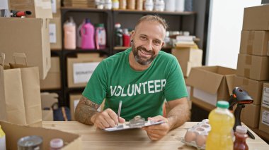 Gülümseyen genç adam, gönüllü olarak hayır kurumunda çalışıyor, masada oturuyor, bağış kutularıyla çevrili, gerekli notları alıyor.