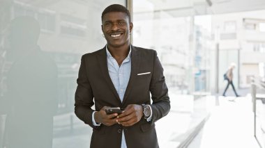 Güneşli bir şehir sokağında binaların arka planında akıllı telefon kullanan şık giyimli yakışıklı Afrikalı bir adam..