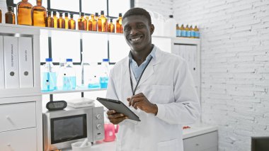 Modern bir laboratuvarda duran laboratuvar önlüklü, gülümseyen Afrikalı adam..