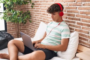 Evde dizüstü bilgisayar kullanan İspanyol bir genç. Yatak odasında oturmuş, düşünceli ve ayık bir ifade takınıyor. Kendinden emin görünüyor. 