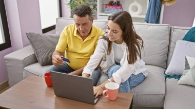 İspanyol bir baba ve gülümseyen bir kız evde online alışveriş yapıyor, dizüstü bilgisayar ve kredi kartıyla oturma odasındaki kanepede oturuyor, birlikte bir fincan espresso içiyorlar.