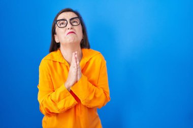 Orta yaşlı İspanyol kadın mavi arka planda gözlük takıyor. Elleriyle yalvarıyor ve dua ediyor. Yüzünde umut ifadesi var. Yalvarıyor. 