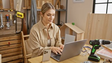 Çekici sarışın kadın marangoz tutkuyla internetteki marangozluk işine dalmış laptopuyla, kapalı atölyesindeki odunların arasında gülümsüyor..
