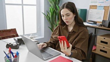 Modern ofis ortamında akıllı telefon ve laptop kullanan odaklanmış genç bir kadın..