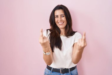 Young brunette woman standing over pink background showing middle finger doing fuck you bad expression, provocation and rude attitude. screaming excited 