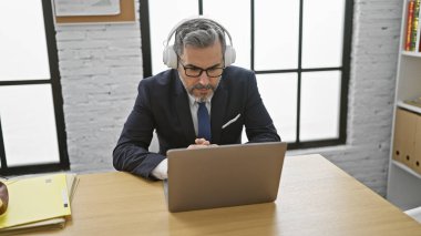 Ciddi İspanyol asıllı genç bir adam, gri saçlı bir iş adamı, iş yerindeki iş yerindeki bilgisayarında ve kulaklıklarıyla başarıya odaklanmış..