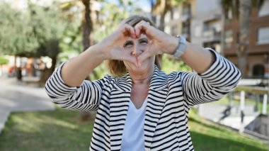 Gülümseyen olgun kadın güneşli parkta kalp şeklinde eller yapıyor.