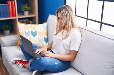Evdeki kanepede dizüstü bilgisayar kullanan genç sarışın kadın.