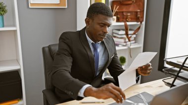 Belgeleri incelerken takım elbise giymiş, özenle modern bir ofiste çalışan odaklanmış Afrikalı bir adam..