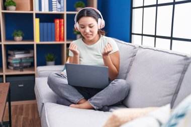 Evde dizüstü bilgisayar kullanan İspanyol genç bir kadın kulaklık takıyor gururla bağırıyor, zaferi kutluyor ve başarıyı kollarını kaldırarak kutluyor. 