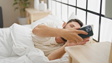 Genç İspanyol adam yatakta akıllı telefon kullanıyor.