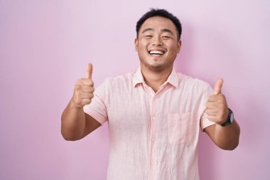 Pembe arka plan başarı işaretinin üzerinde duran Çinli genç adam elleriyle olumlu jest yapıyor, gülümsüyor ve mutlu. Neşeli ifade ve kazanan jest. 