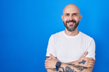 Mavi arka planda dikilen dövmeli İspanyol adam kollarını kavuşturup kameraya bakarken gülümsüyor. pozitif kişi. 