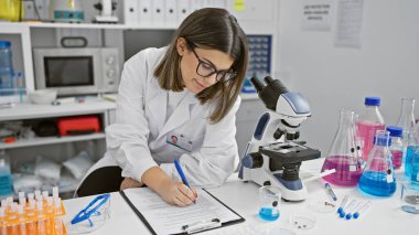 Genç İspanyol bilim kadını bilimsel ekipman ve renkli kimyasallarla dolu bir laboratuvarda notlar alıyor..