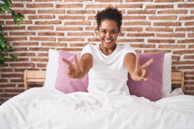 Afro-Amerikan kadın zafer jesti yapıyor. Yatak odasında oturuyor.