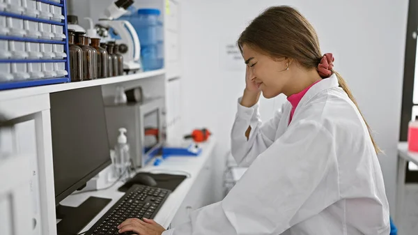 Struggling with stress at work, young, beautiful hispanic woman scientist in pain, suffering a headache while typing on her computer in a lab.