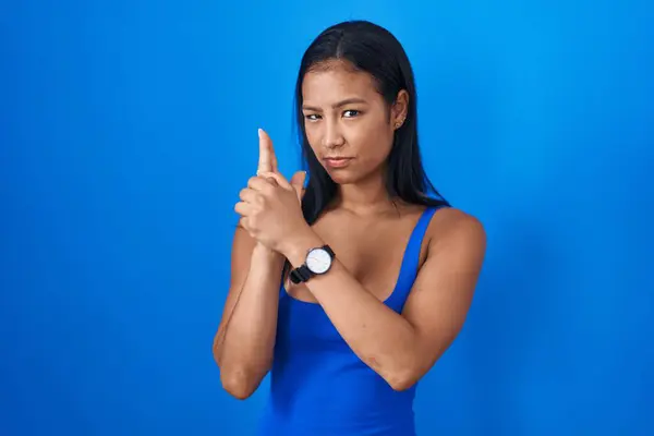Hispanic woman standing over blue background holding symbolic gun with hand gesture, playing killing shooting weapons, angry face