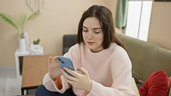 A young woman in casual attire focusing on her smartphone in a cozy, well-lit living room setting.