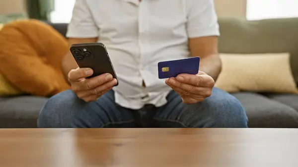 A middle-aged man sits at home with a smartphone and credit card, potentially shopping online or managing finances.