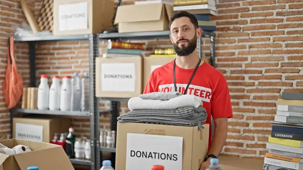 Young hispanic man volunteer holding donation package with clothes at charity center