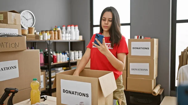 Hardworking young hispanic woman volunteer, beautifully checking clothes donations in charity center package, using her smartphone.