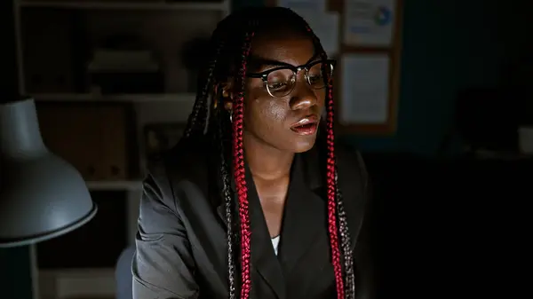 Focused african american woman boss governs her business empire night and day, navigating success from her workplace desk, mastering the internet on her office computer screen